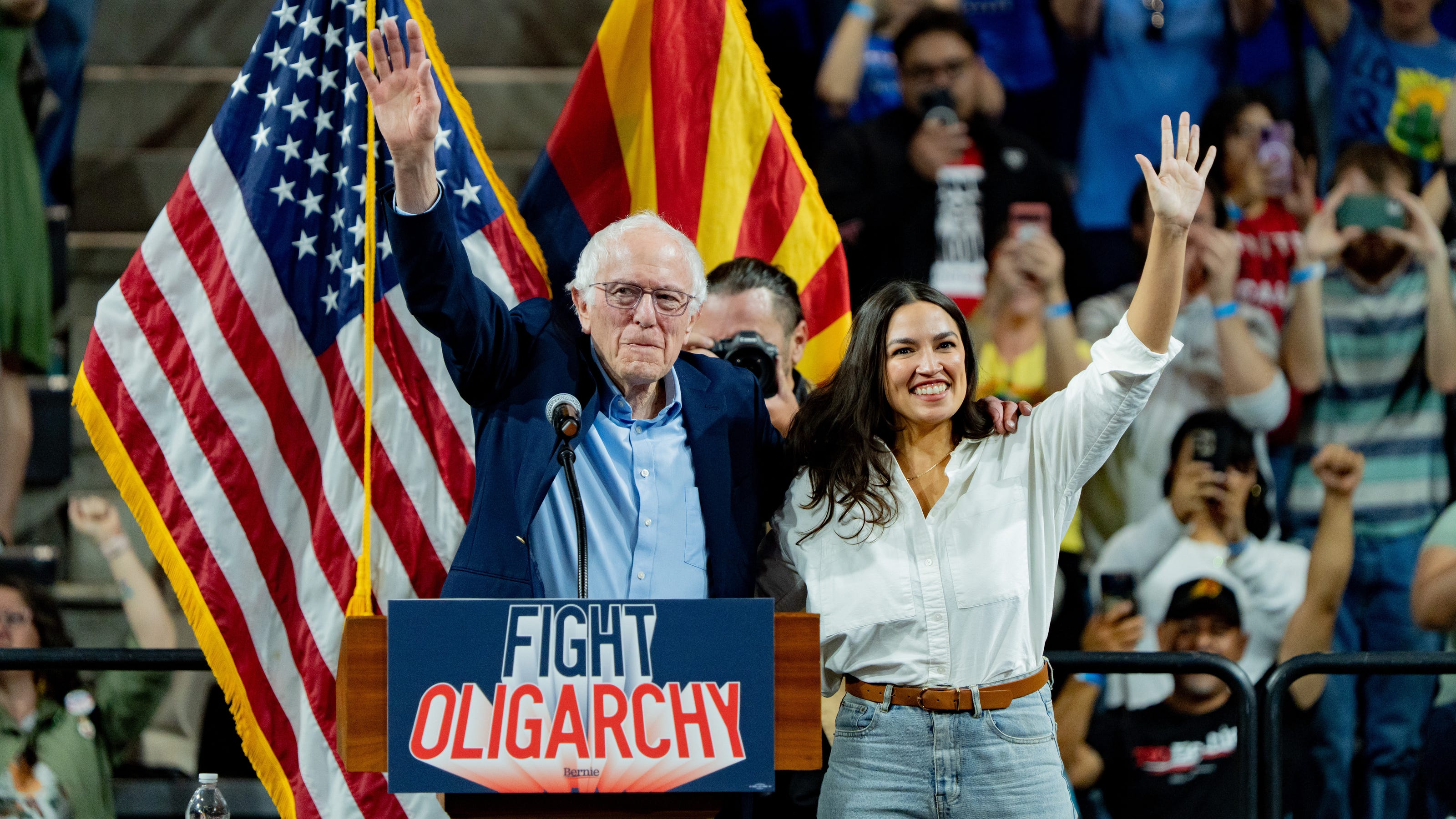 Image of Bernie Sanders and Alexandria Ocasio-Cortez waving at a political rally, behind a podium with a sign reading “fight oligarchy.”
