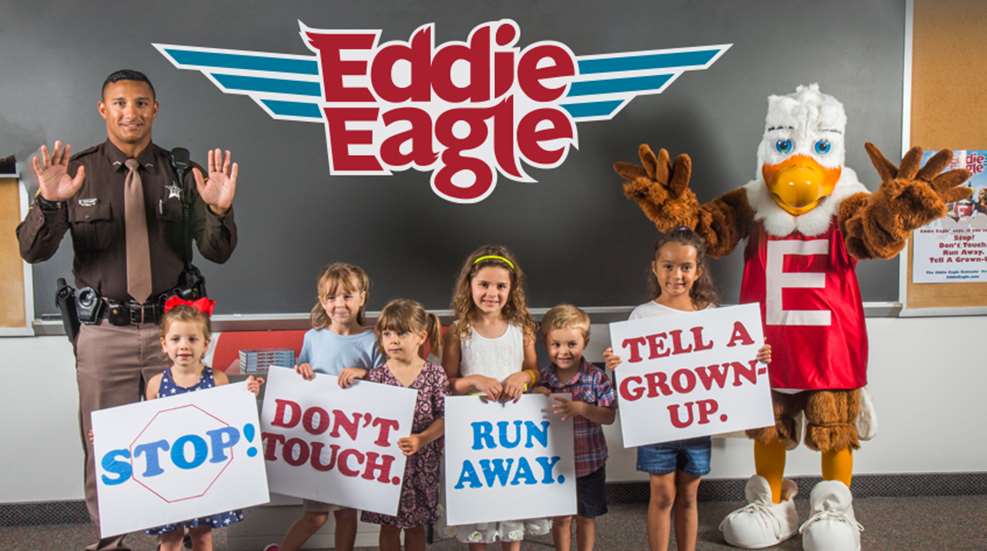 Photograph of schoolchildren posing with a police officer and a mascot character named "Eddie Eagle," holding signs reading "stop," "run away," and "don't touch."