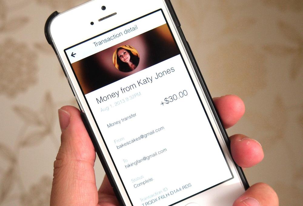 Image of a hand holding an iPhone, showing a transaction in the Google Wallet app for transferring money to a friend.