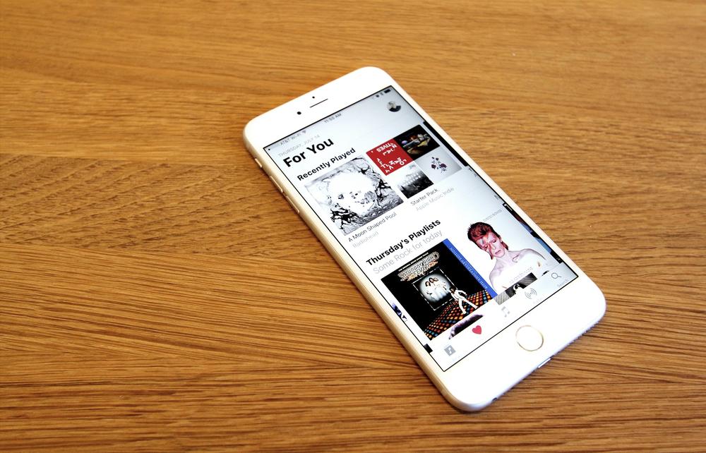 An image of an iPhone 6s resting on a wooden table, running the Apple Music app in iOS 10. The home screen of Apple Music shows the new, bolder headers that are central to the new iOS 10 design aesthetic.