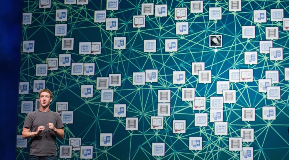 An image of Facebook CEO Mark Zuckerberg presenting at a conference, with a large visualization of the Facebook social graph behind him.